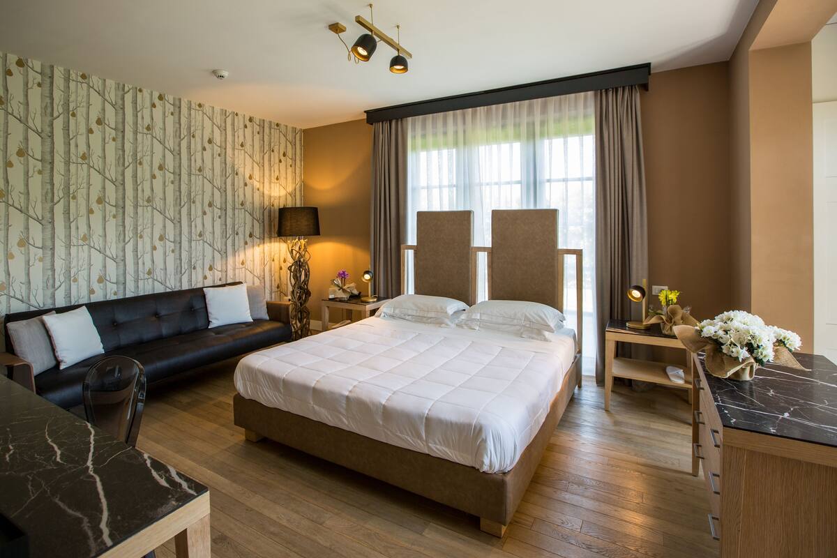 Una habitación de hotel moderna con una cama grande, un sofá, un escritorio y una silla. Cuenta con una ventana con cortinas, una lámpara y un jarrón con flores.