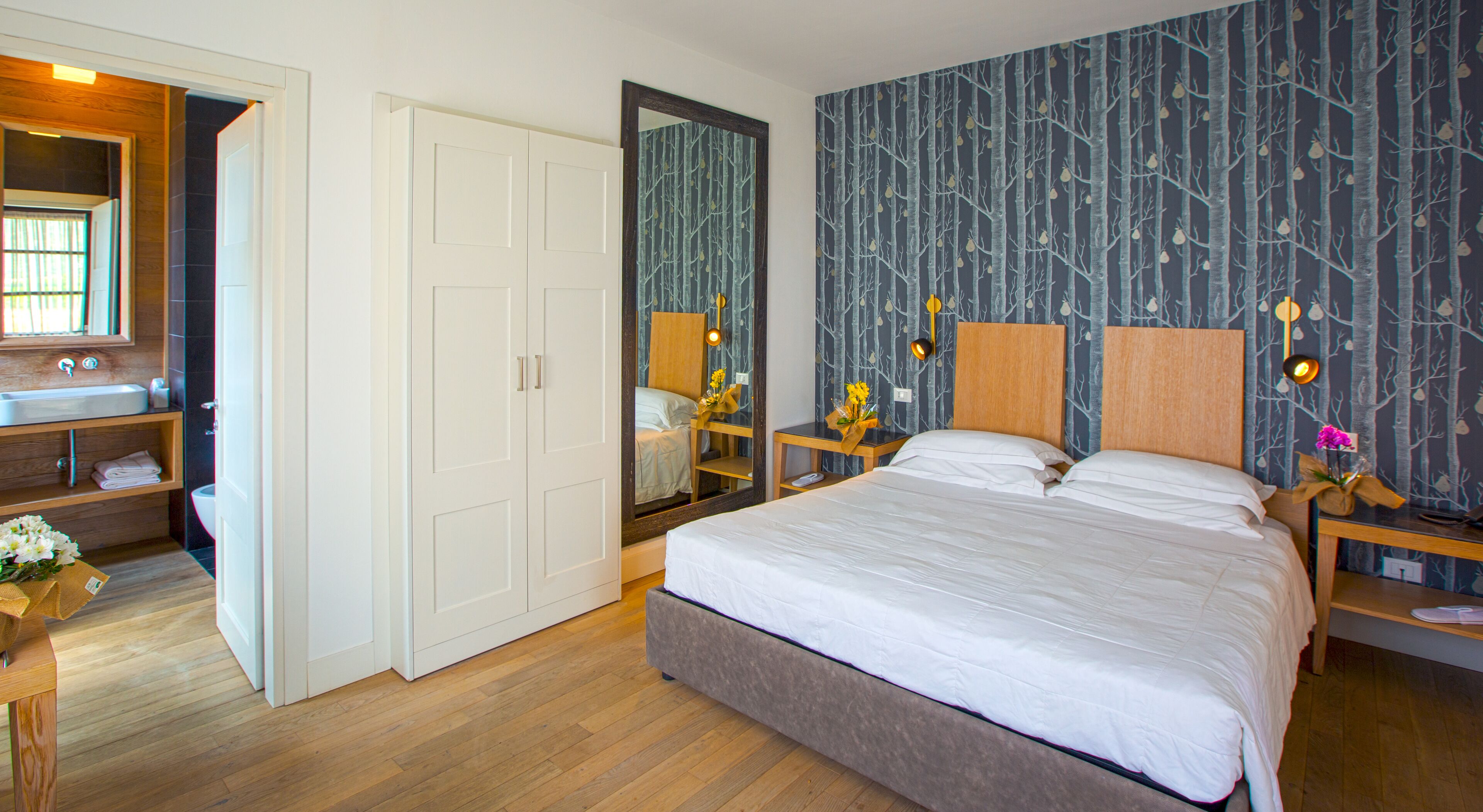 Een slaapkamer met een groot bed, een houten nachtkastje, een spiegel, een dressoir, en een badkamer zichtbaar door een geopende deur.
