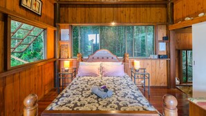 A wooden cabin bedroom with a large bed, two bedside tables, and a window with a view of the forest. - The Epiphyte Bed & Breakfast (Cow Bay)