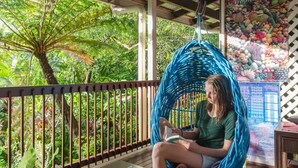 A person sitting in a blue woven hammock chair on a wooden deck with a view of lush greenery and a tropical fruit poster in the background. - The Epiphyte Bed & Breakfast (Cow Bay)
