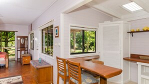 A room with a wooden dining table, two chairs, a bench, a small wooden cabinet, and a window with a view of greenery. - The Epiphyte Bed & Breakfast (Cow Bay)