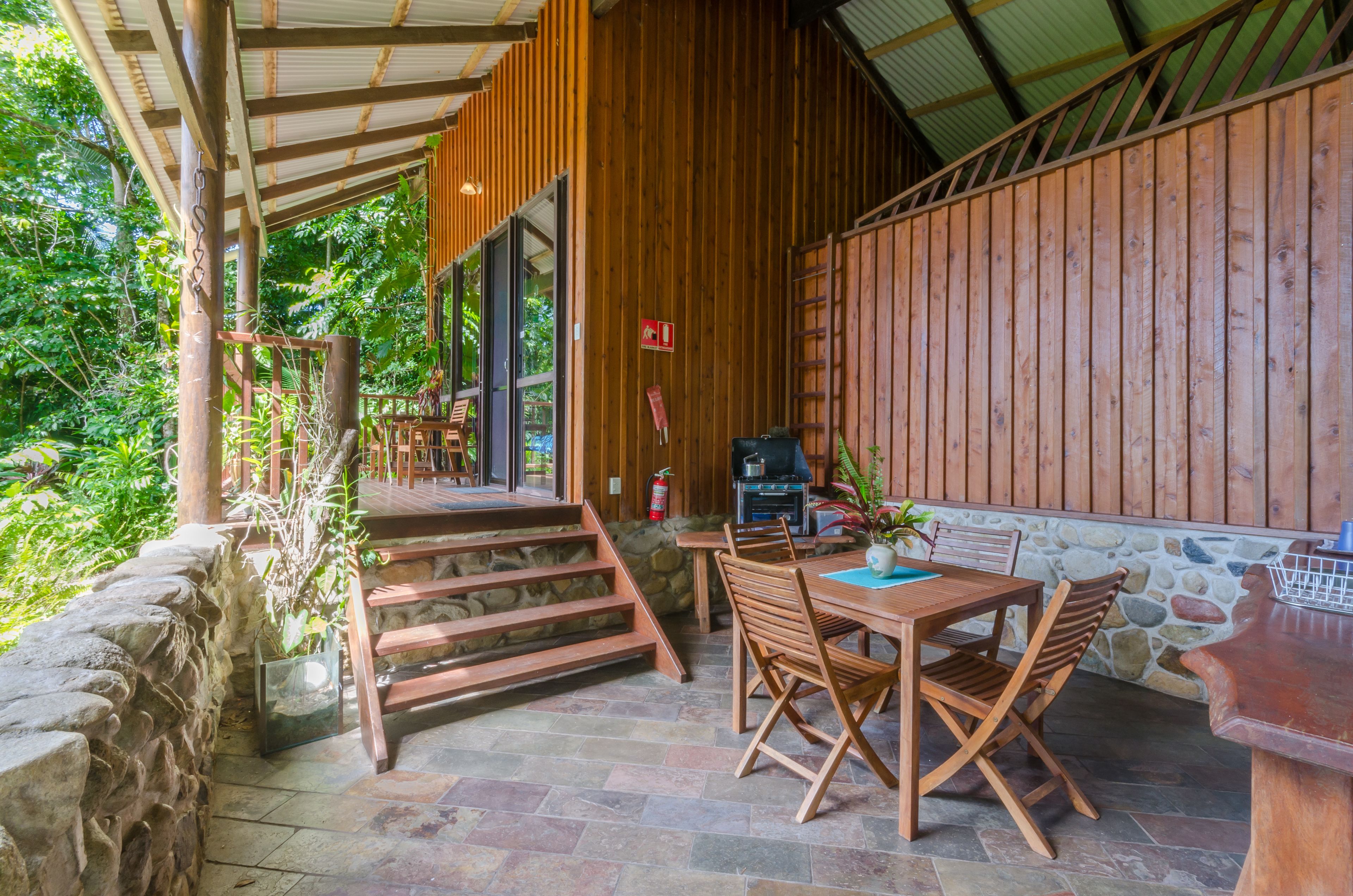 Une terrasse extérieure couverte, aménagée avec du mobilier en bois, un mur en pierre et offrant une vue sur la verdure environnante.