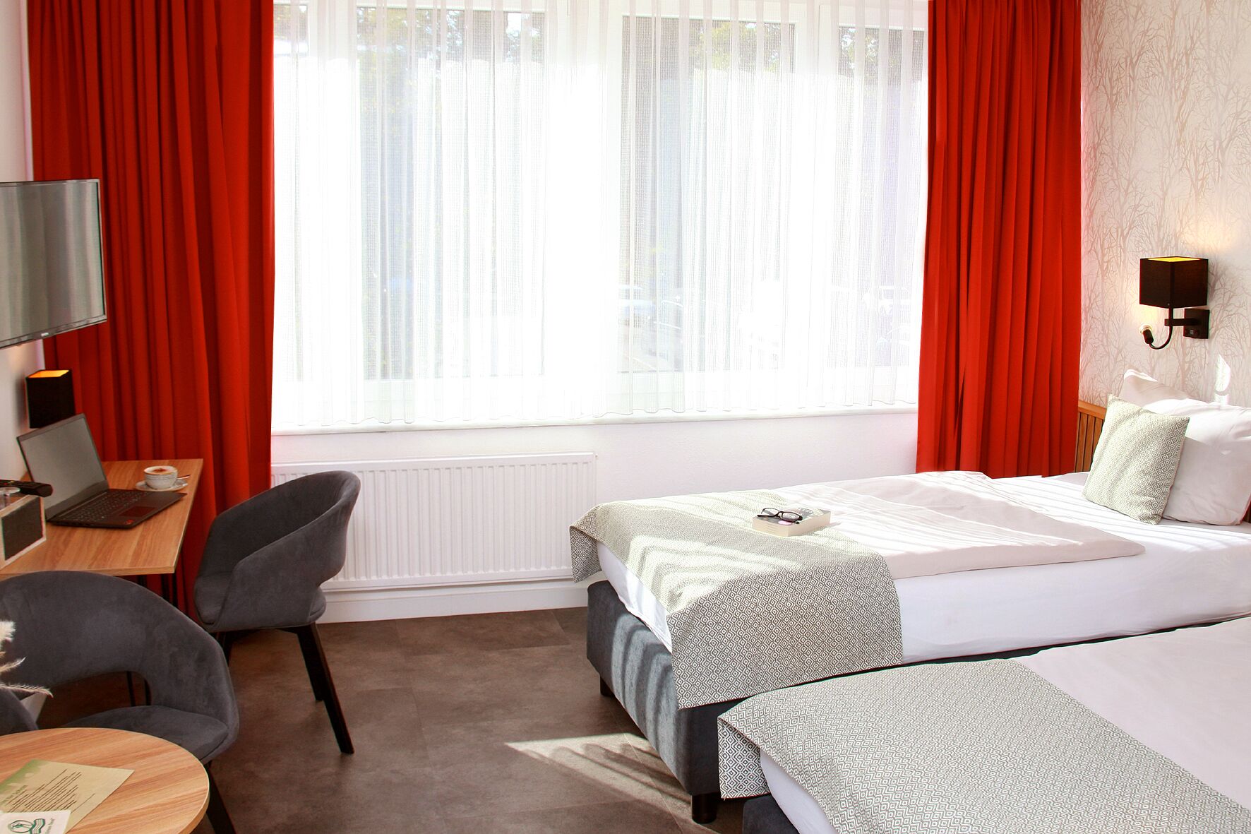 A hotel room with a bed, a desk with a laptop, a chair, and a window with red curtains.