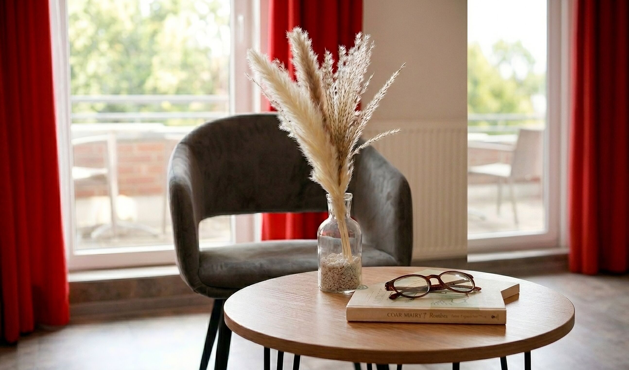 A round wooden table with a book and glasses, a vase with dried plants, and a chair in a room with red curtains and a view through a window.