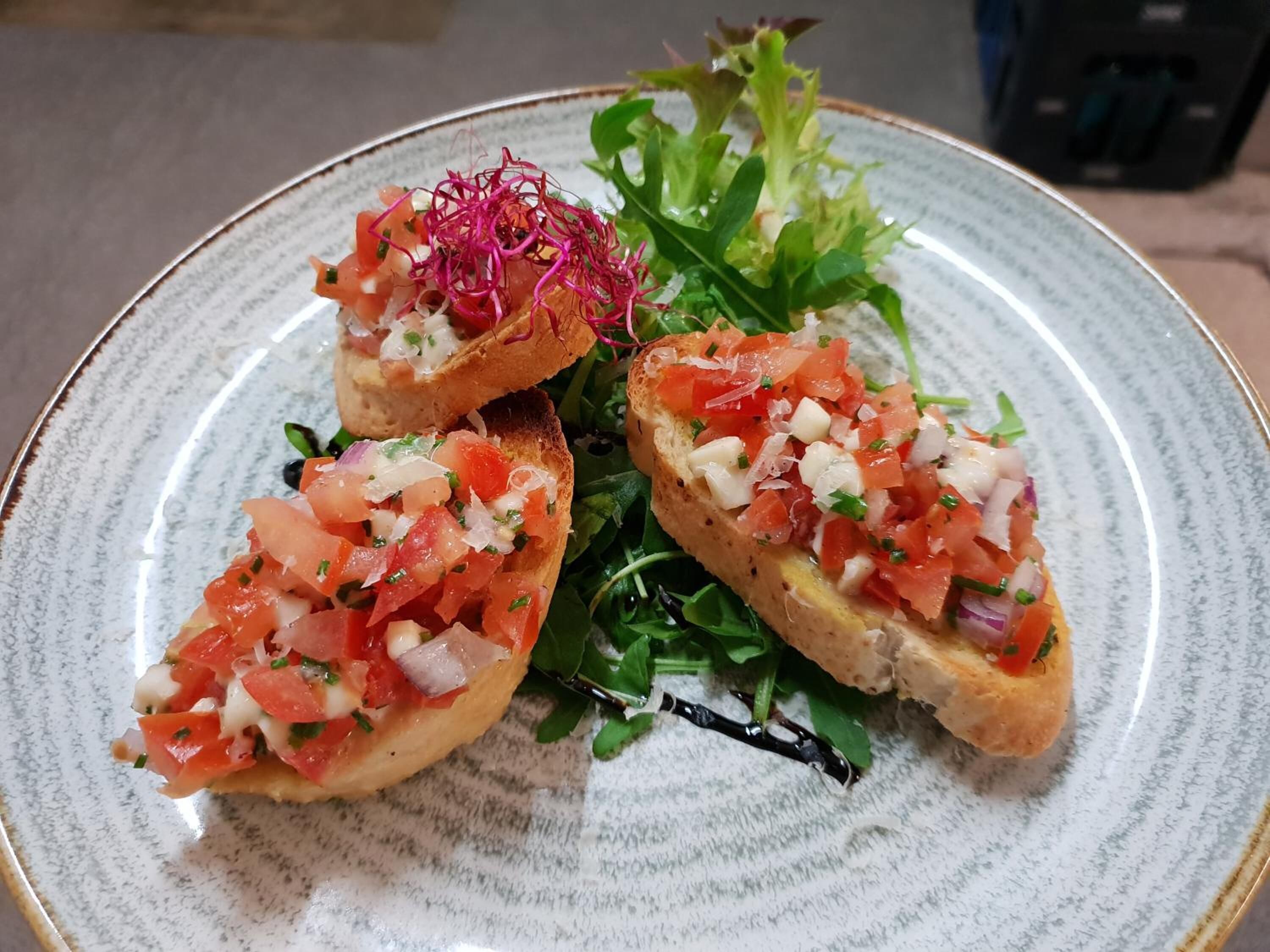 Un plato de bruschetta cubierto de tomates y cebollas picados, adornado con microgreens y una ramita de flores moradas.