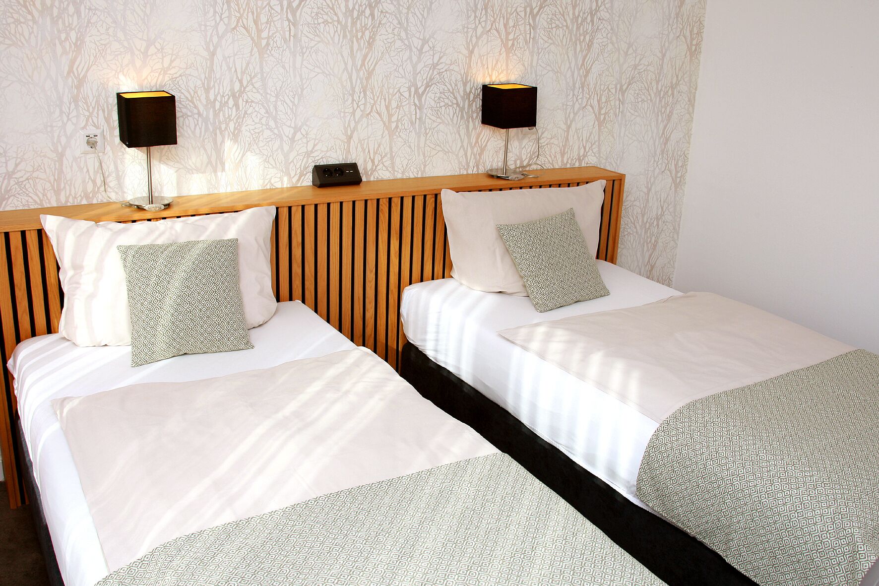 A hotel room with two beds, each with a wooden headboard, white bedding, and patterned throw pillows. There are two wall-mounted lamps on either side of the headboards.