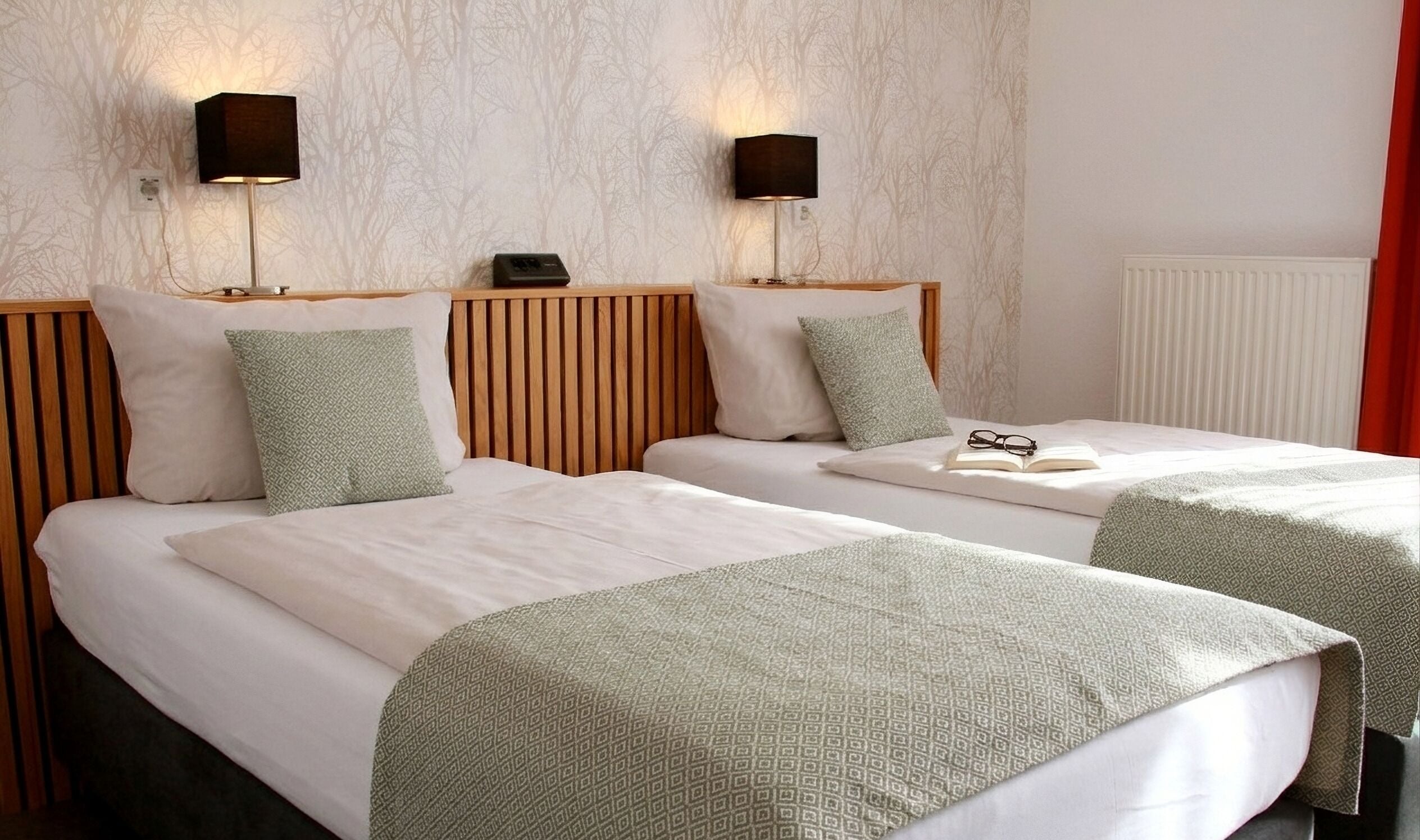A hotel room with two beds, each with a wooden headboard, white bedding, and green patterned throw pillows. There are wall-mounted lamps on either side of the beds.