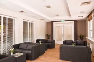 A modern lounge area with dark wicker furniture, large windows with blinds, and a wooden floor.