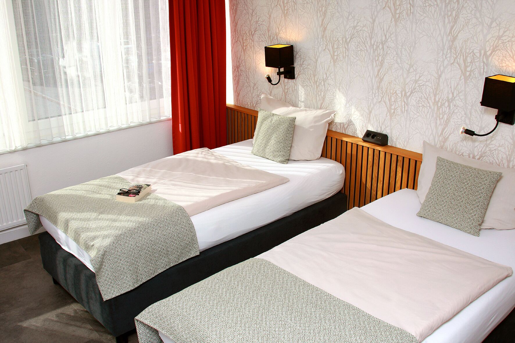 Habitación de hotel con dos camas, una ventana con cortinas traslúcidas, una cortina roja, ropa de cama estampada y luces fijadas a la pared.