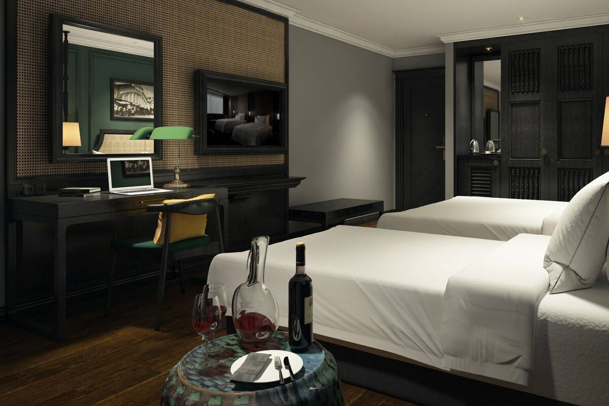 Habitación de hotel con cama, escritorio con una computadora portátil, silla, mesa redonda con una botella y vasos, y un espejo.