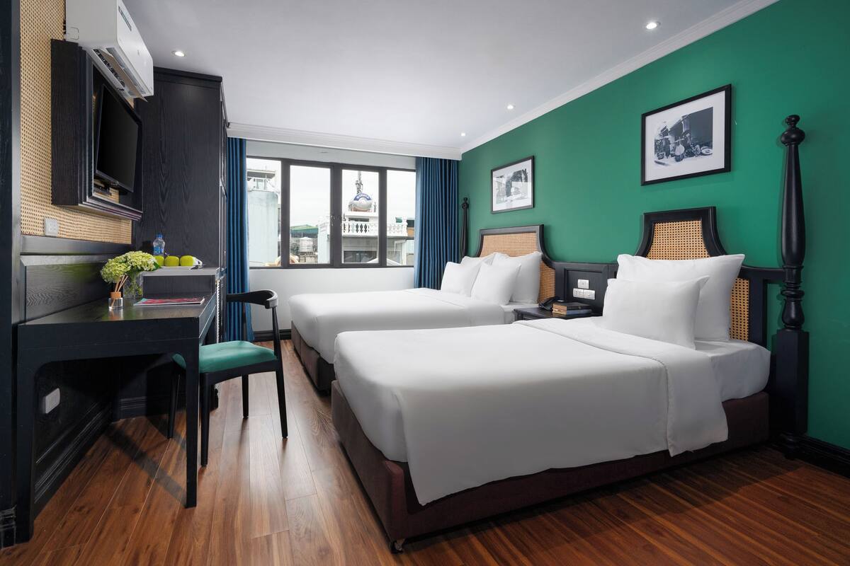 Habitación de hotel con dos camas, un escritorio con silla, televisión y ventana con vistas a edificios.