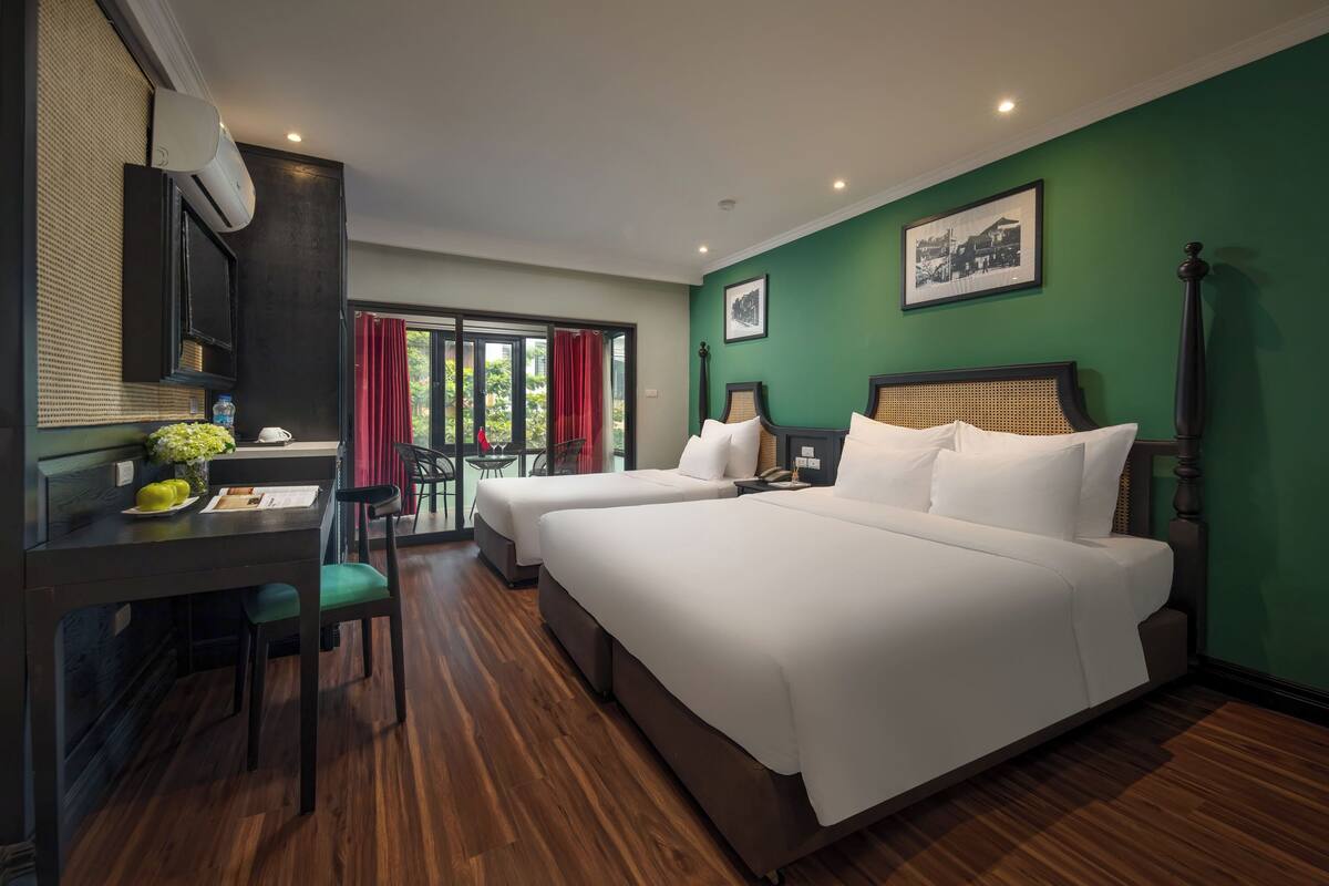 Una habitación de hotel con dos camas, un escritorio con una silla, un ventanal con cortinas rojas y una pared verde con un cuadro enmarcado.