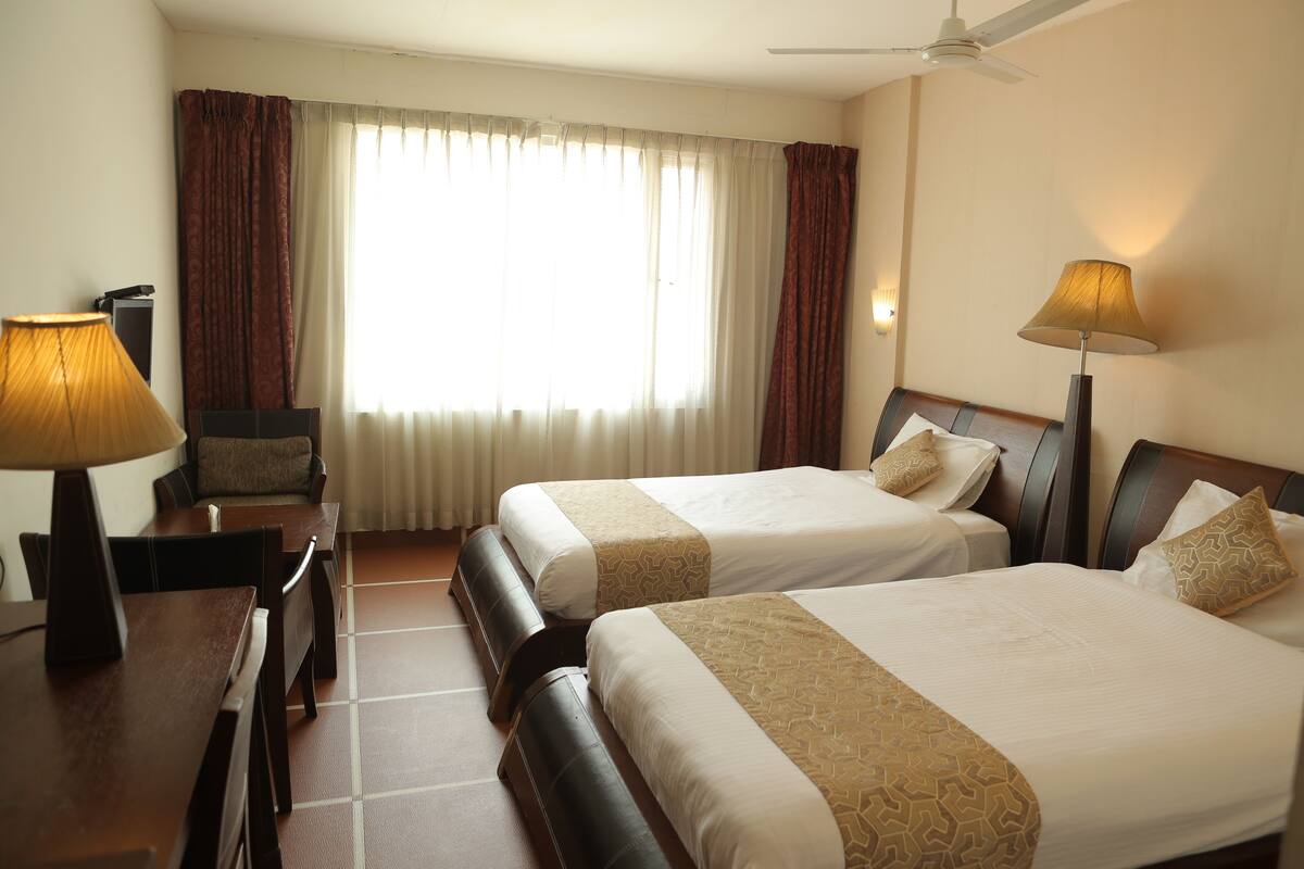 A hotel room with two beds, a desk, a chair, a lamp, and a ceiling fan.