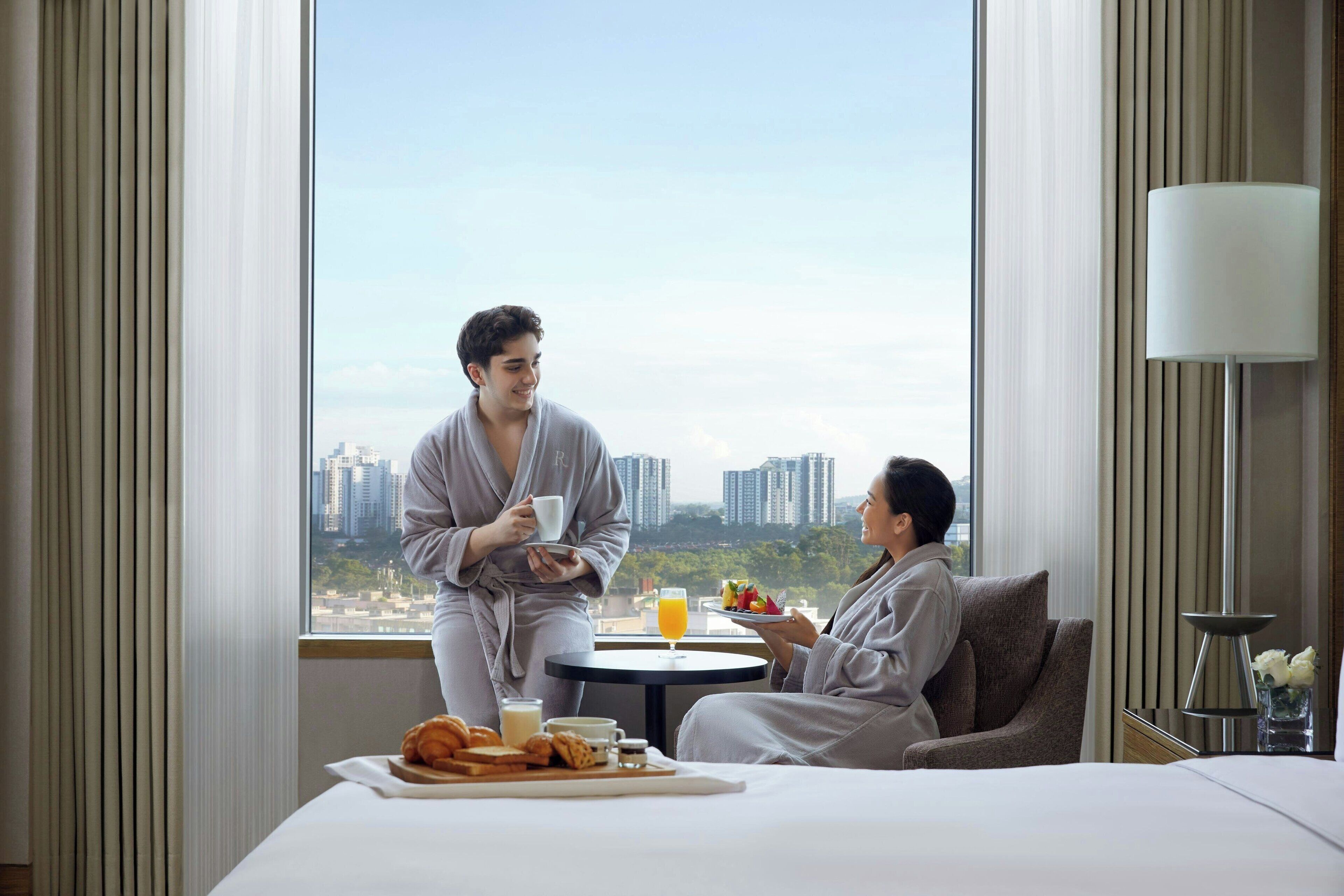 Een hotelkamer met een groot raam, een bed, een nachtkastje met een lamp en een ontbijtkarretje met toast en sap.