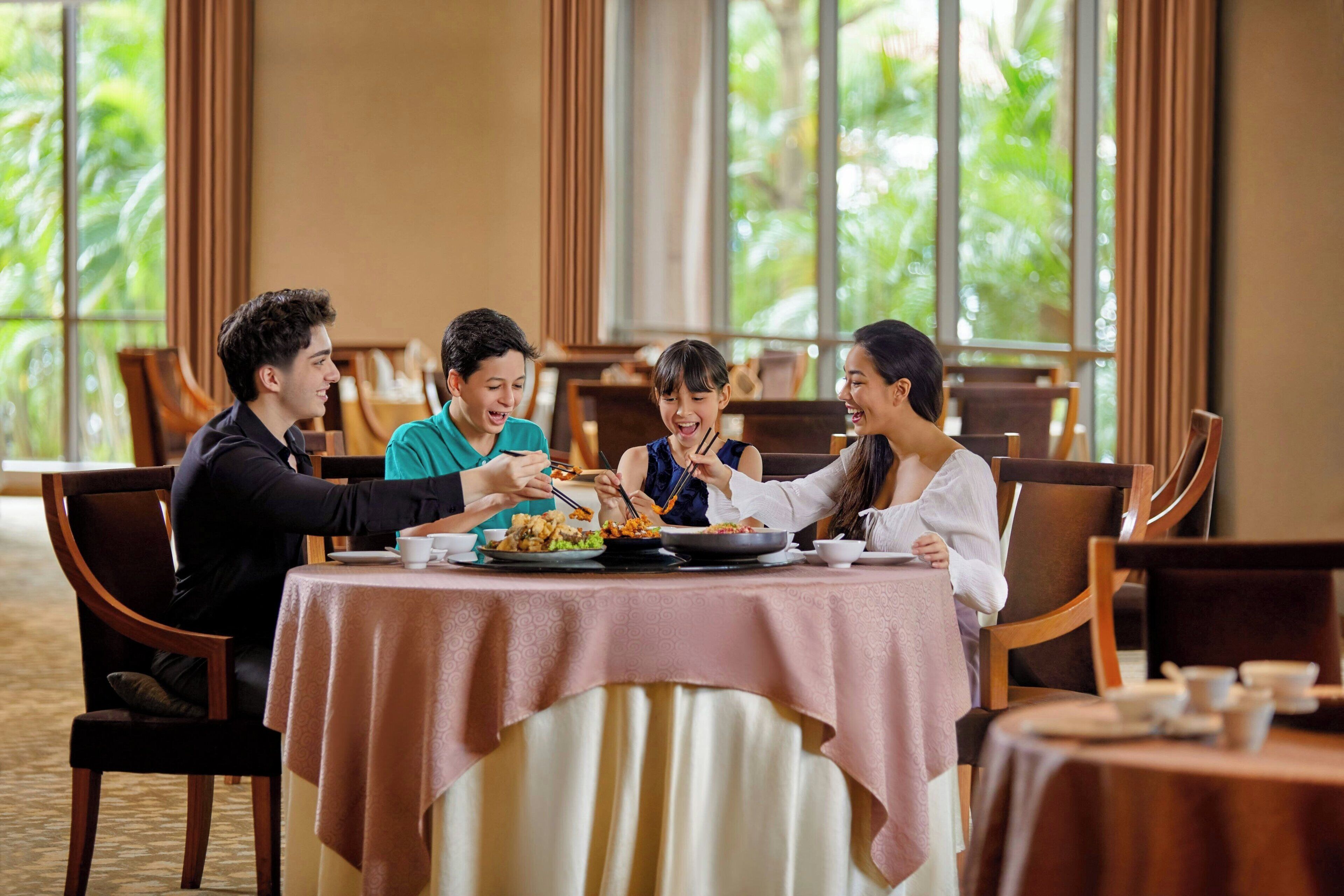 A group of people dining at a table with a tablecloth, surrounded by chairs and a window with a view of greenery.