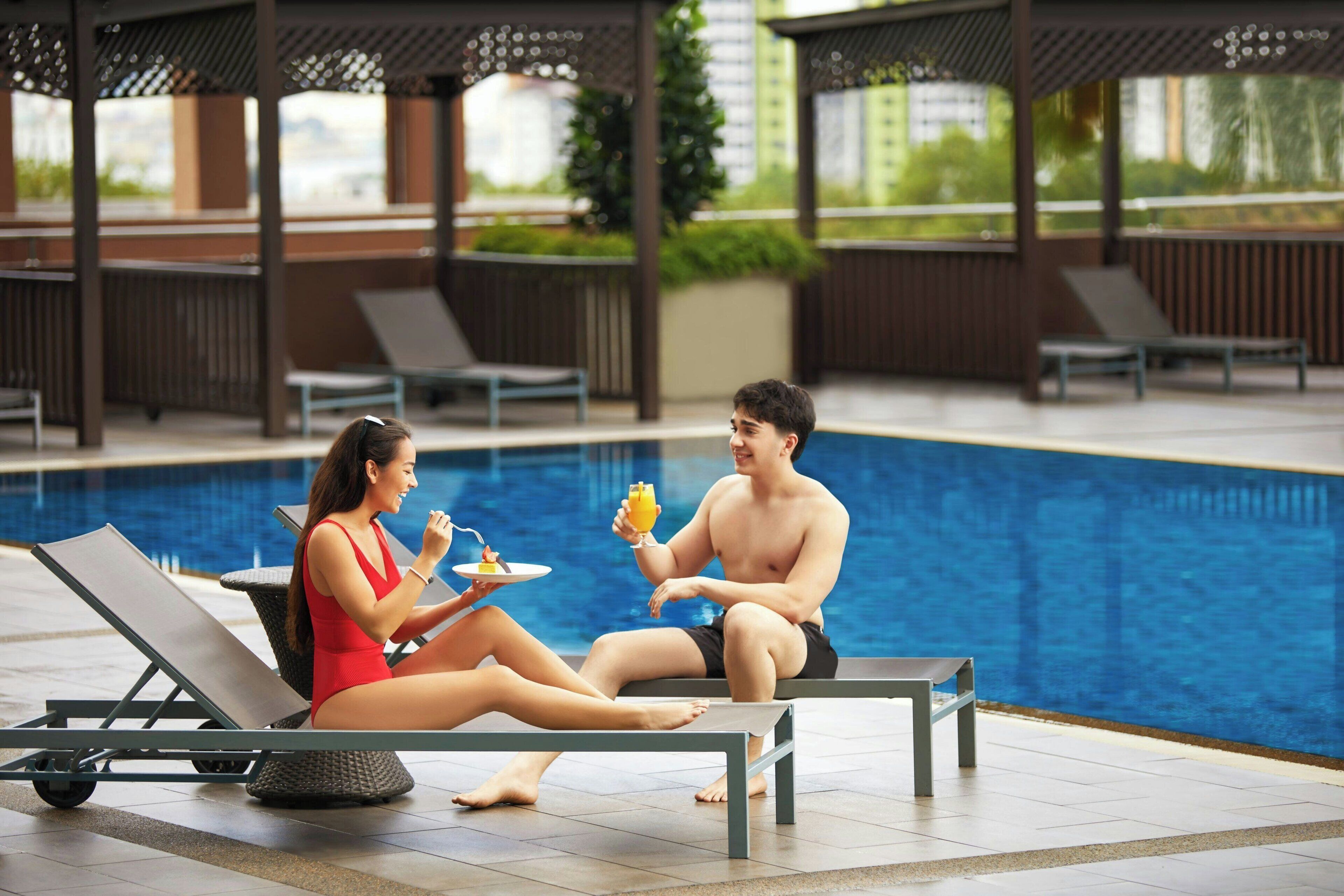 A couple relaxing by a poolside lounge chair with a drink tray, overlooking a swimming pool.