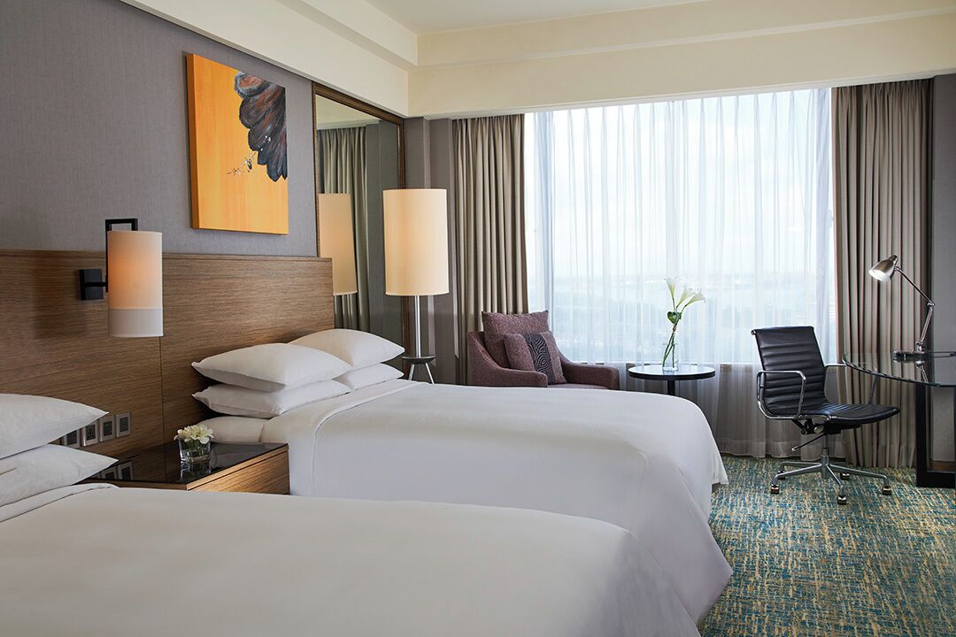 A hotel room with two beds, a large window with curtains, a painting on the wall, and a small table with a vase of flowers.