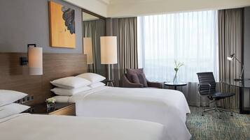 Habitación de hotel con dos camas, una zona de estar con una silla y una mesa, un ventanal grande con cortinas y un cuadro en la pared.