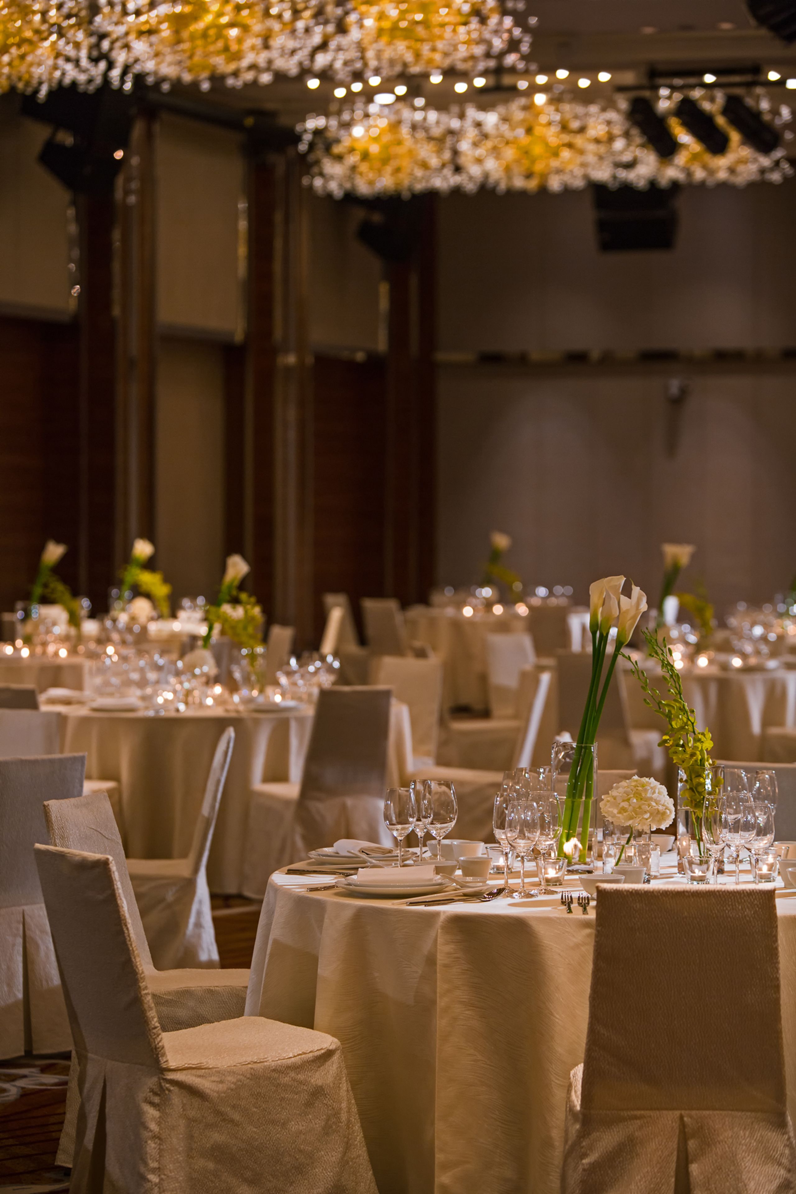 Une salle de banquet avec des tables rondes dressées pour le repas, chacune étant recouverte d’une nappe blanche et entourée de chaises.