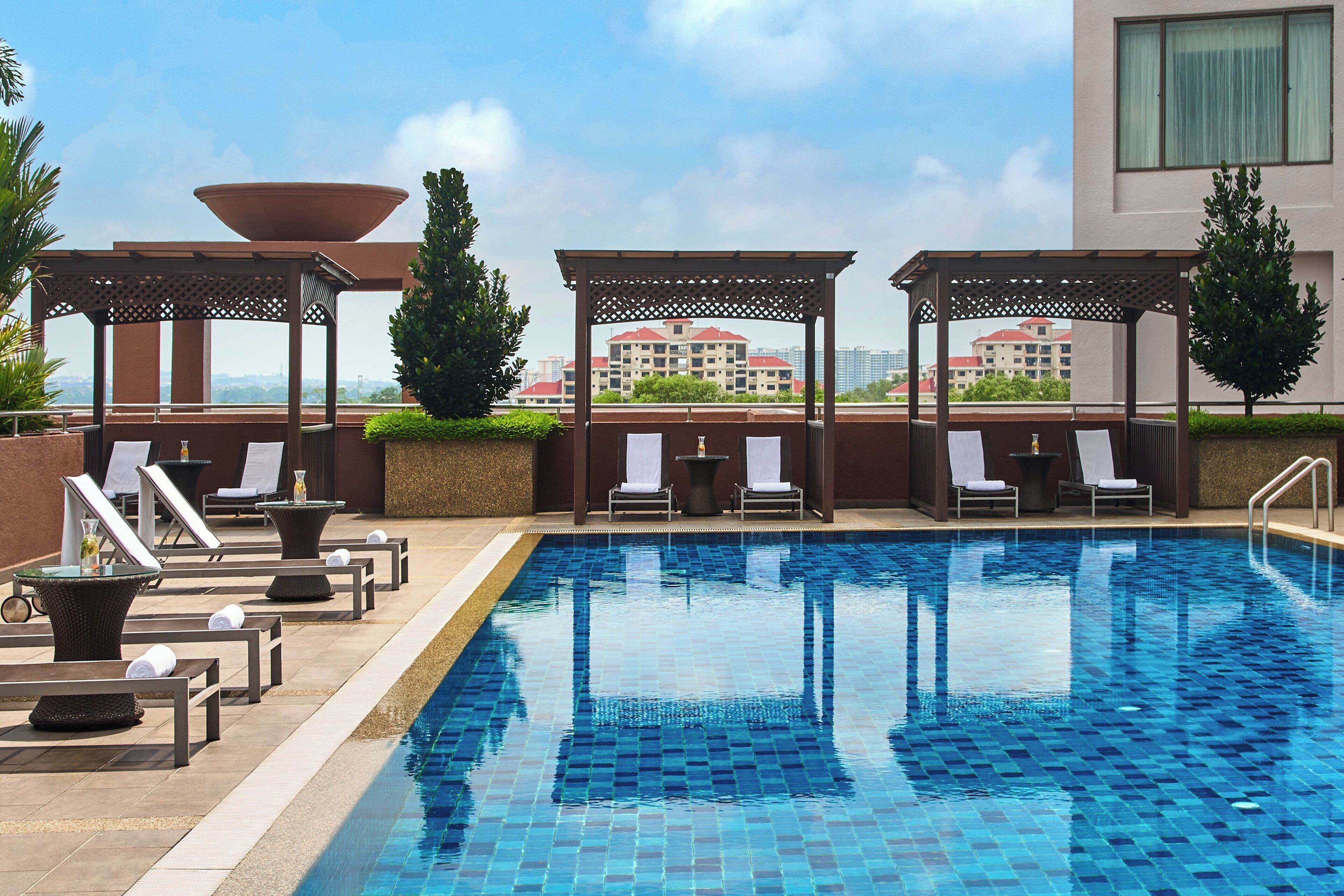 A rooftop pool area with lounge chairs and a pergola, overlooking a cityscape.