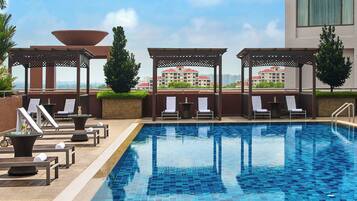 A rooftop pool area with lounge chairs and a pergola, overlooking a cityscape.