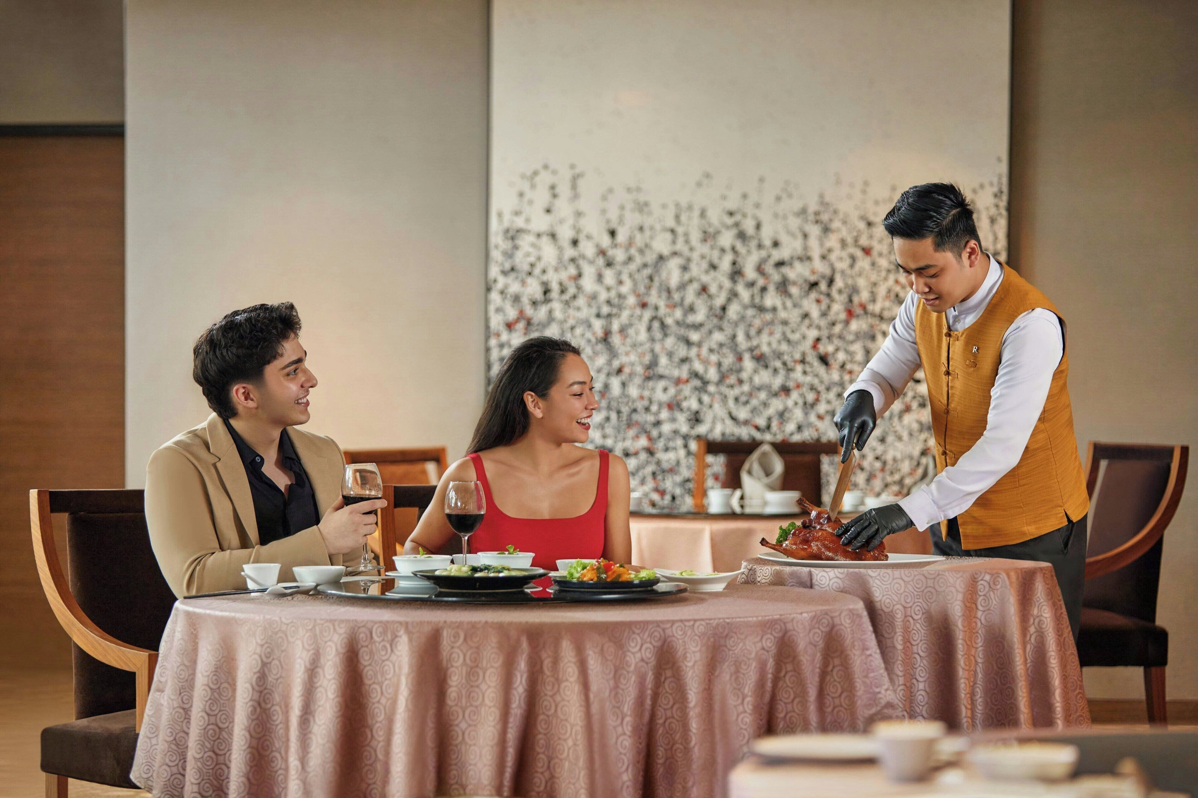 A dining table with a patterned tablecloth, a waiter serving food, and two diners.