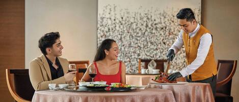 A dining table with a patterned tablecloth, a waiter serving food, and two diners.