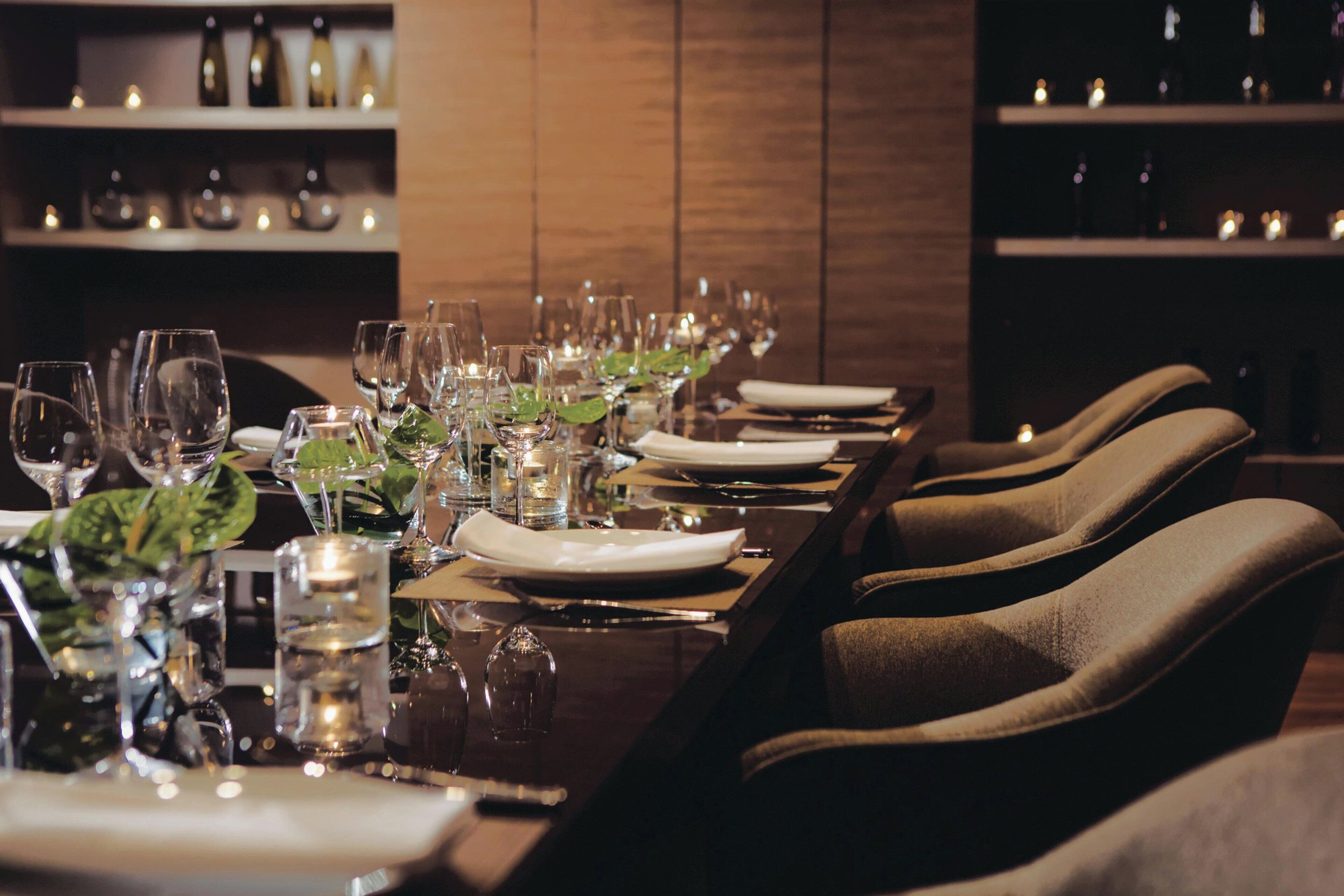 Una larga mesa de comedor dispuesta con copas de vino, platos y velas, flanqueada por sillas tapizadas.