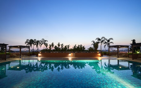 A swimming pool with tiled flooring, surrounded by palm trees and wooden gazebos.