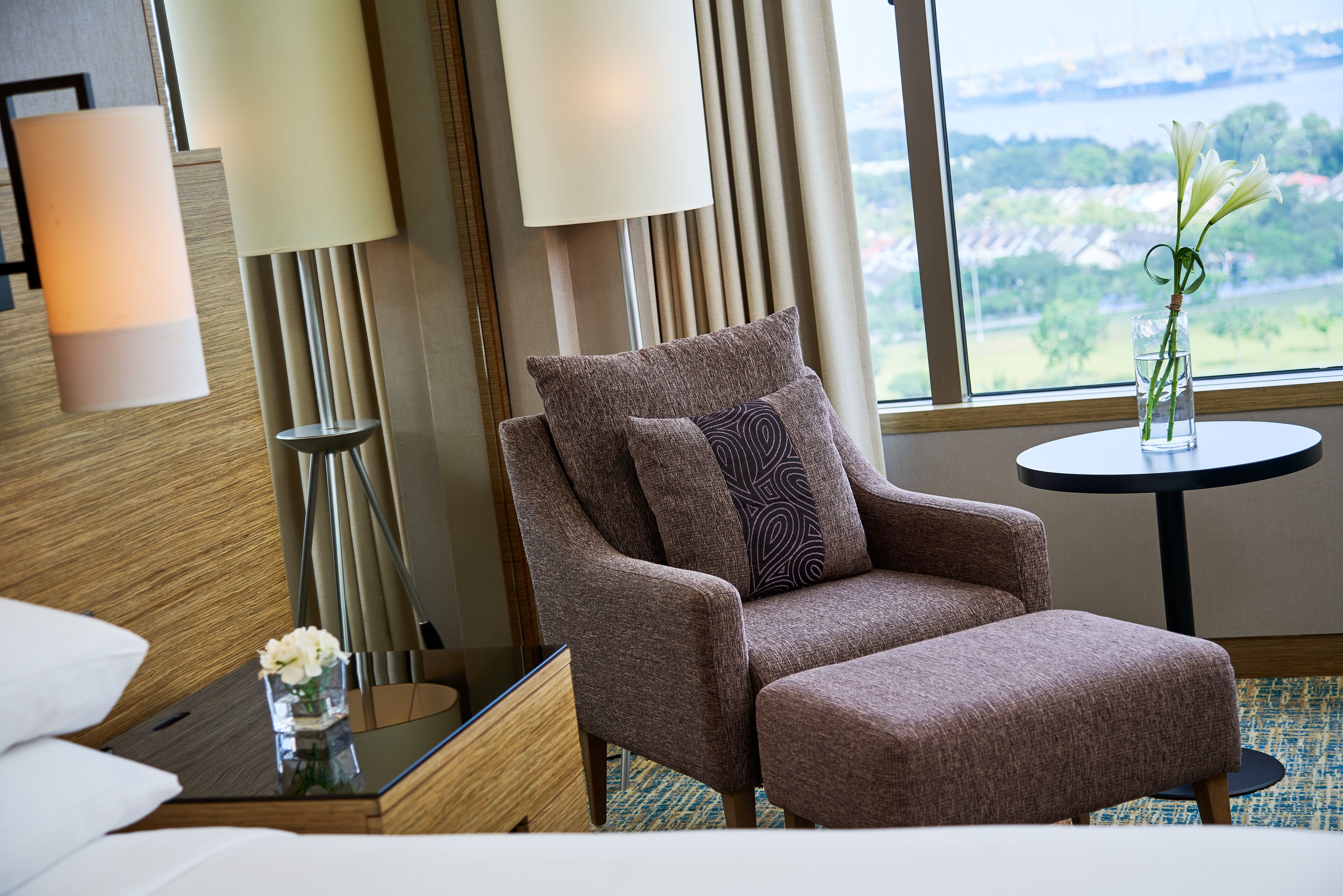 A modern hotel room with a large window, a brown armchair, a small round table, and a vase with flowers.