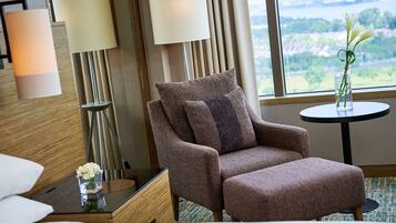 A modern hotel room with a large window, a brown armchair, a small round table, and a vase with flowers.