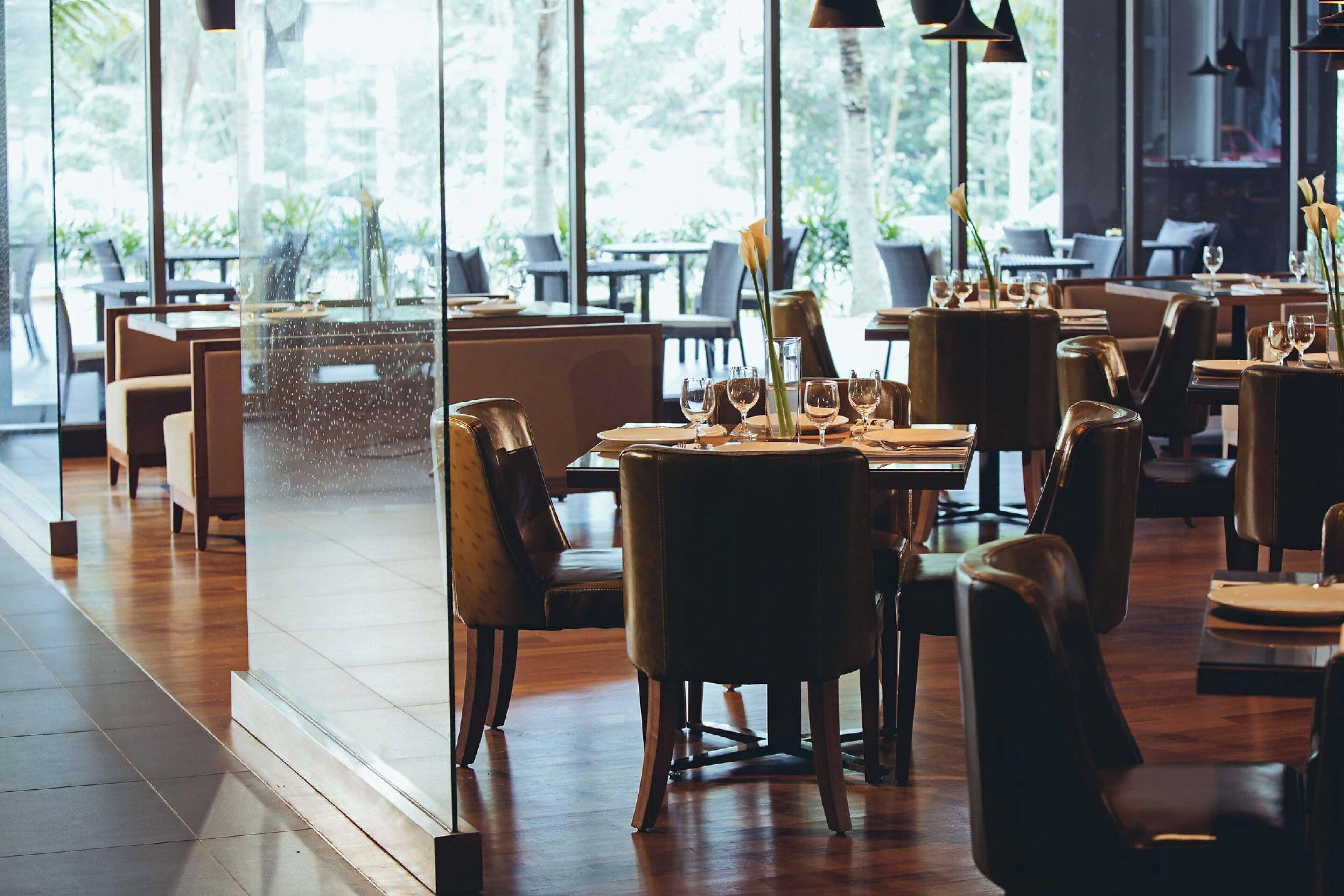 A restaurant with a modern interior, featuring wooden floors and large windows offering a view of the outdoors. Tables are set with wine glasses and plates, and there are chairs arranged around them.