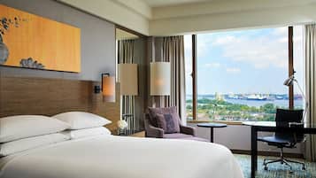 Ein Hotelzimmer mit einem großen Bett, einem Sessel, einem Schreibtisch und Blick auf eine Stadtlandschaft durch große Fenster.