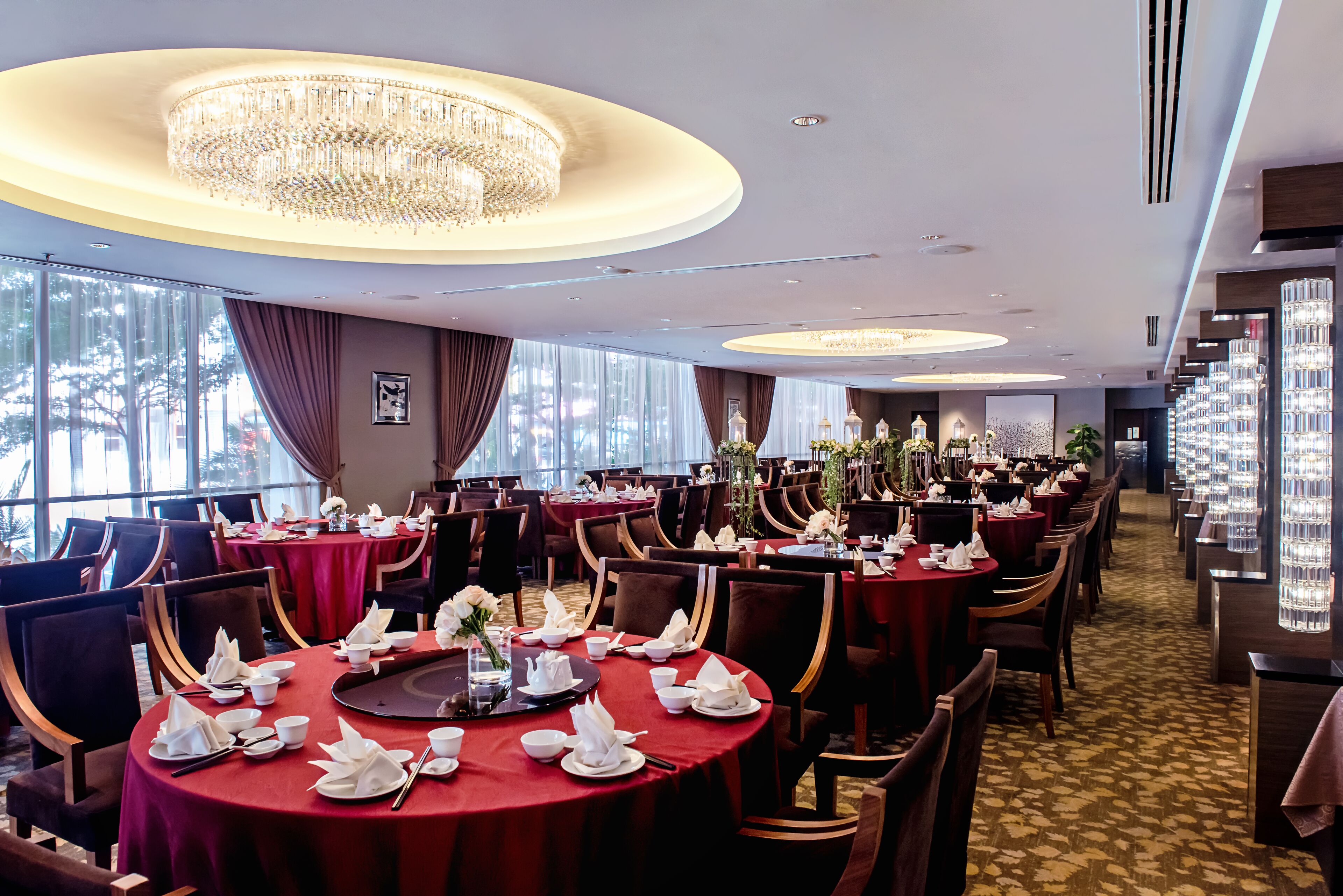 A hotel dining area with multiple round tables set for dining, large chandeliers, and a view of trees through large windows.