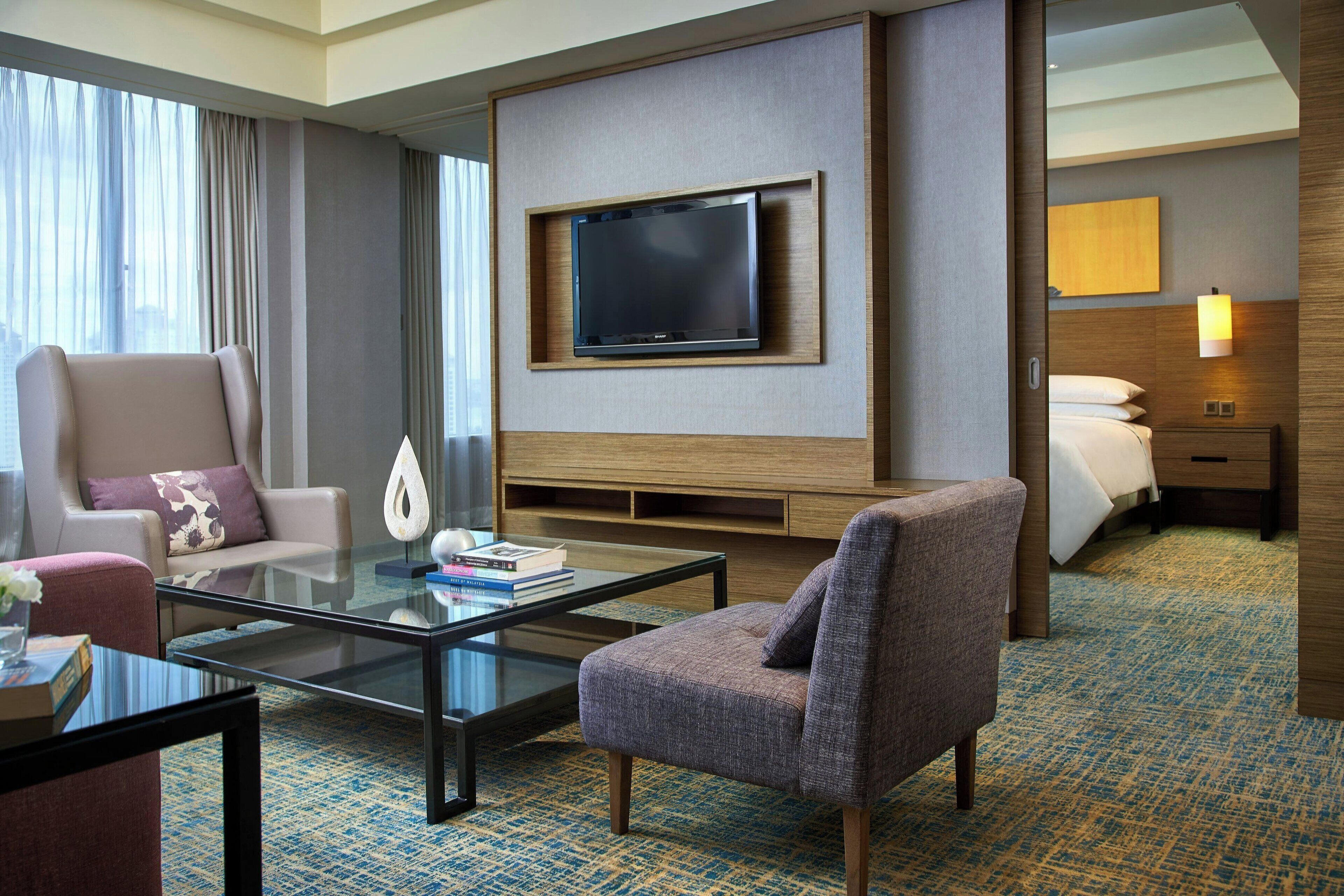 A hotel room with a large flat-screen TV mounted on the wall, a glass coffee table, a comfortable armchair, a sofa, a bed with white linens, and a bedside table with a lamp.