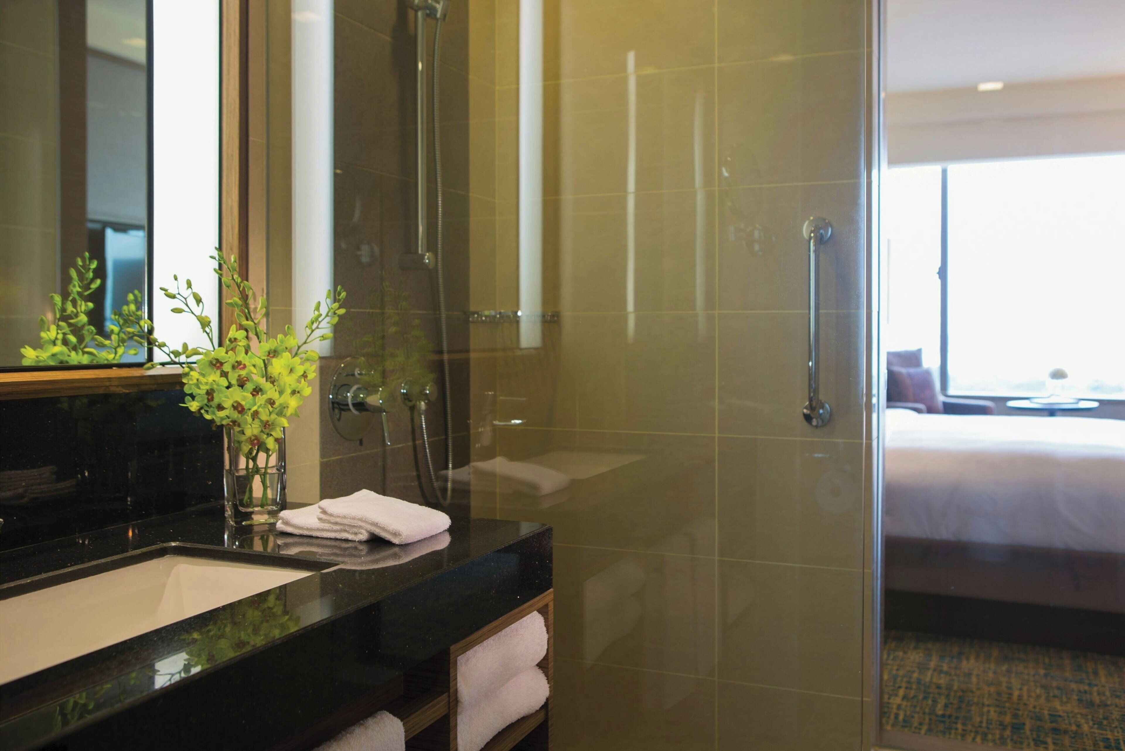 A modern bathroom with a glass shower enclosure, a black countertop, and a visible bed in the adjacent room.
