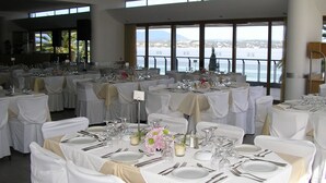 Un salón de banquetes con mesas dispuestas para un evento formal, amplias ventanas con vista a una masa de agua y un letrero de salida de emergencia.