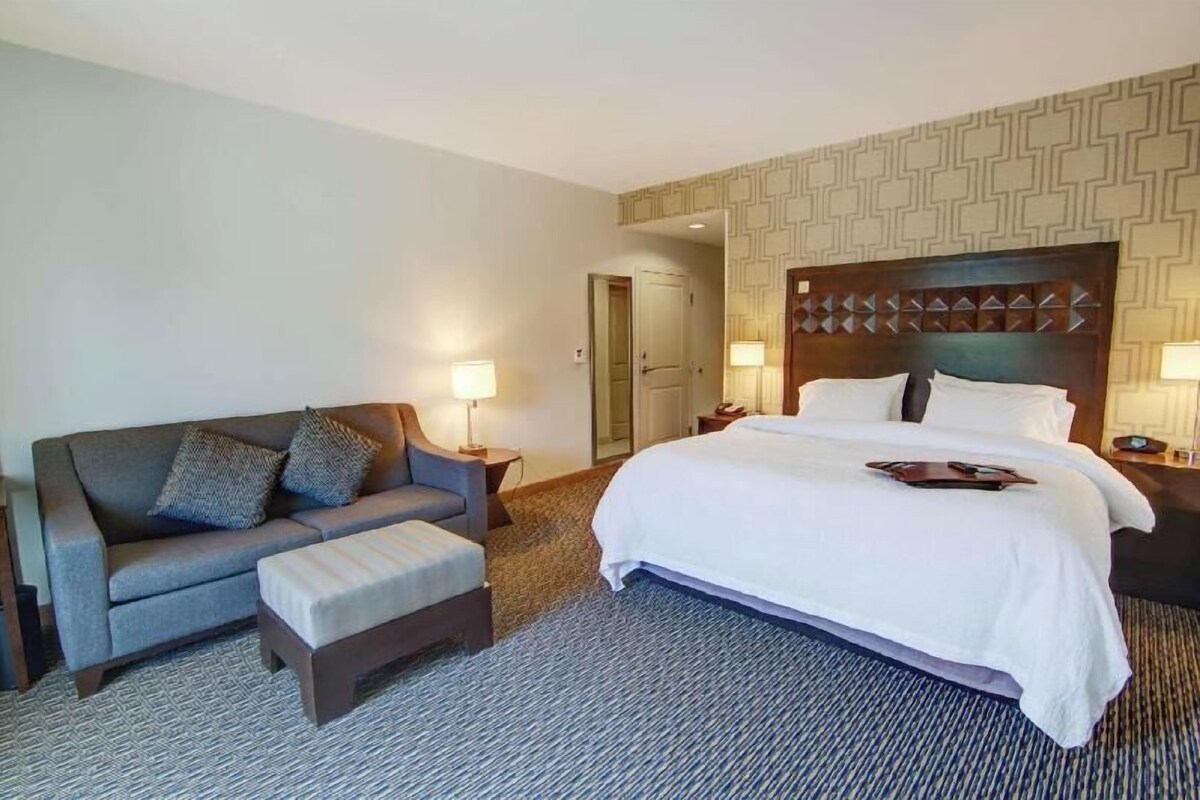 A hotel room with a bed, a sofa, an ottoman, two bedside tables, and a door.