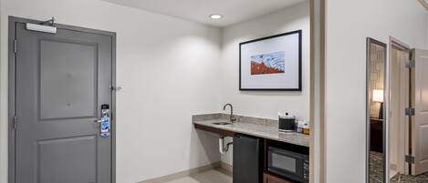 Habitación de hotel compacta con kitchenette, una puerta gris, un cuadro enmarcado en la pared y una alfombra estampada.