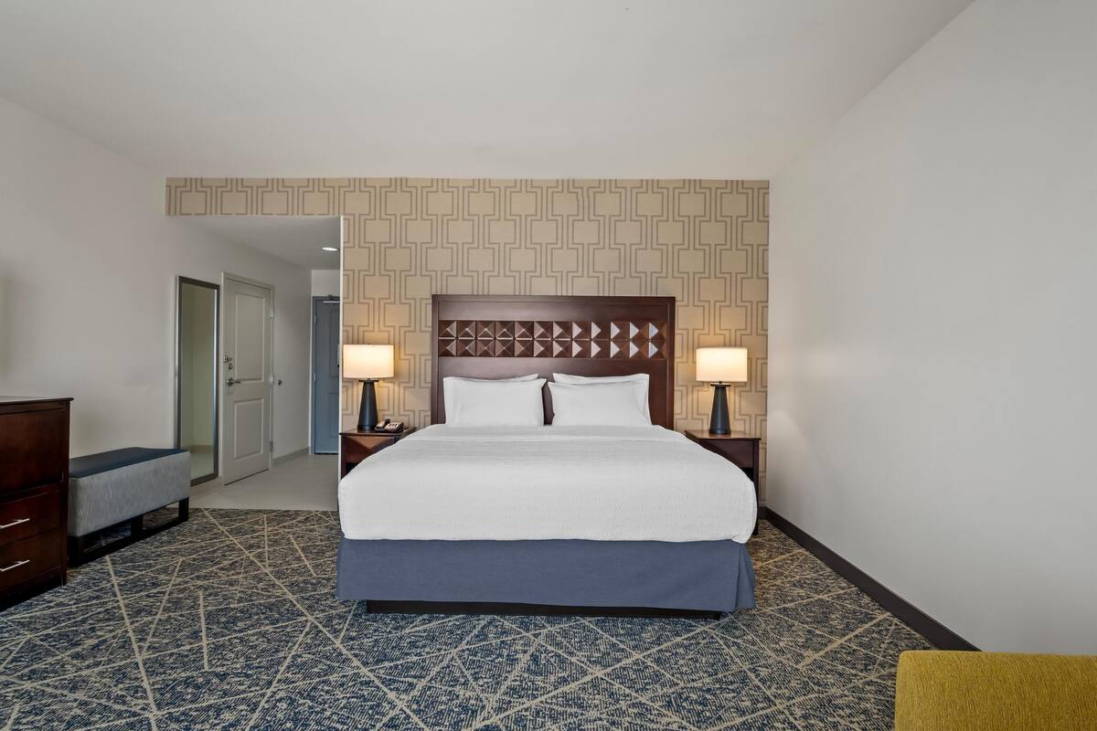 A hotel room with a large bed, two bedside tables with lamps, a wooden headboard, patterned carpet, and a door leading to another room.