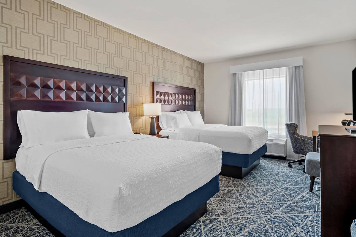 A hotel room with two beds, a desk, a chair, and a large window.