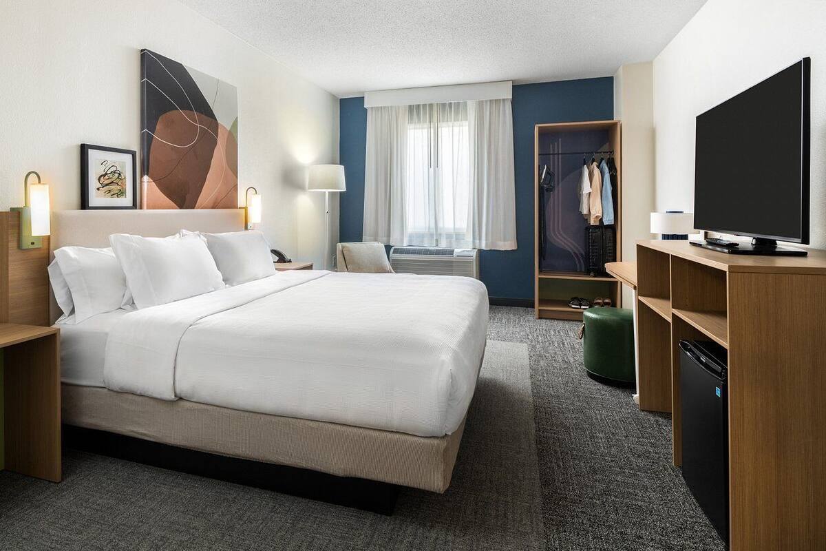 A hotel room with a large bed, a television, a wardrobe, and a desk with a lamp.