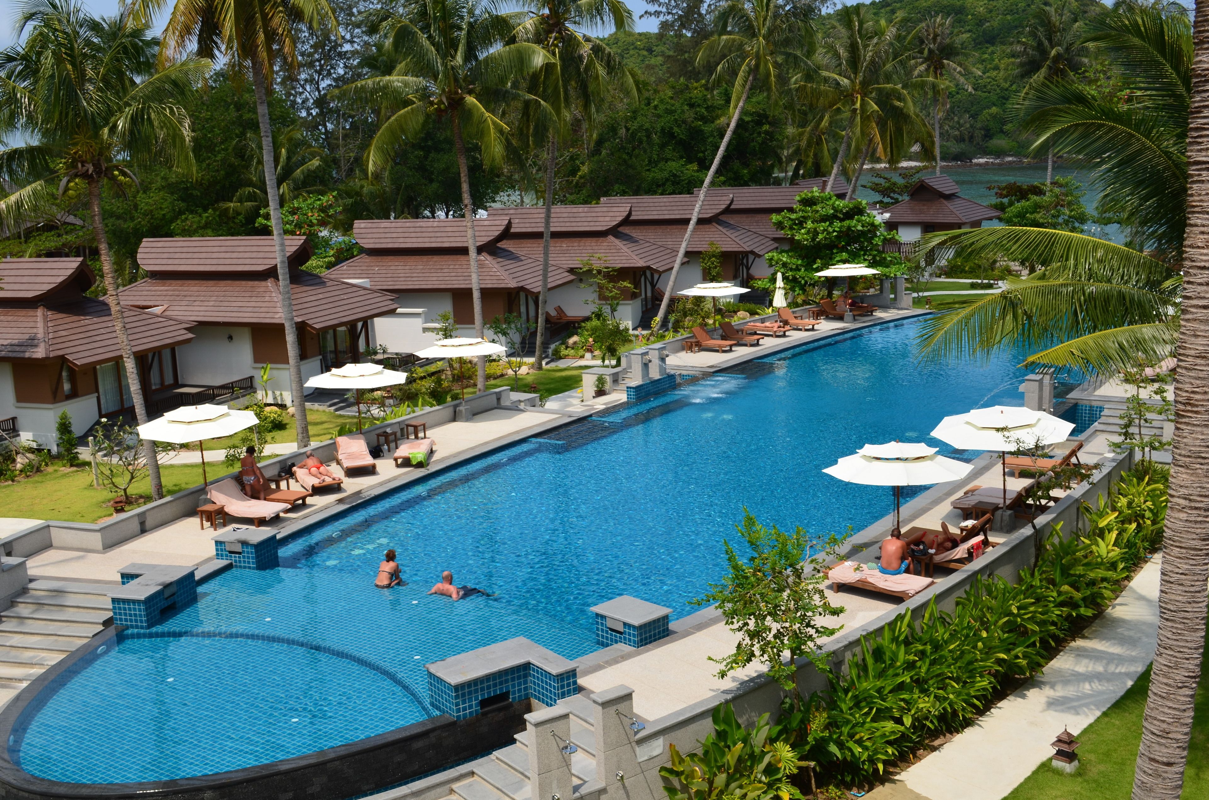 A swimming pool with lounge chairs and umbrellas, surrounded by palm trees and resort buildings.
