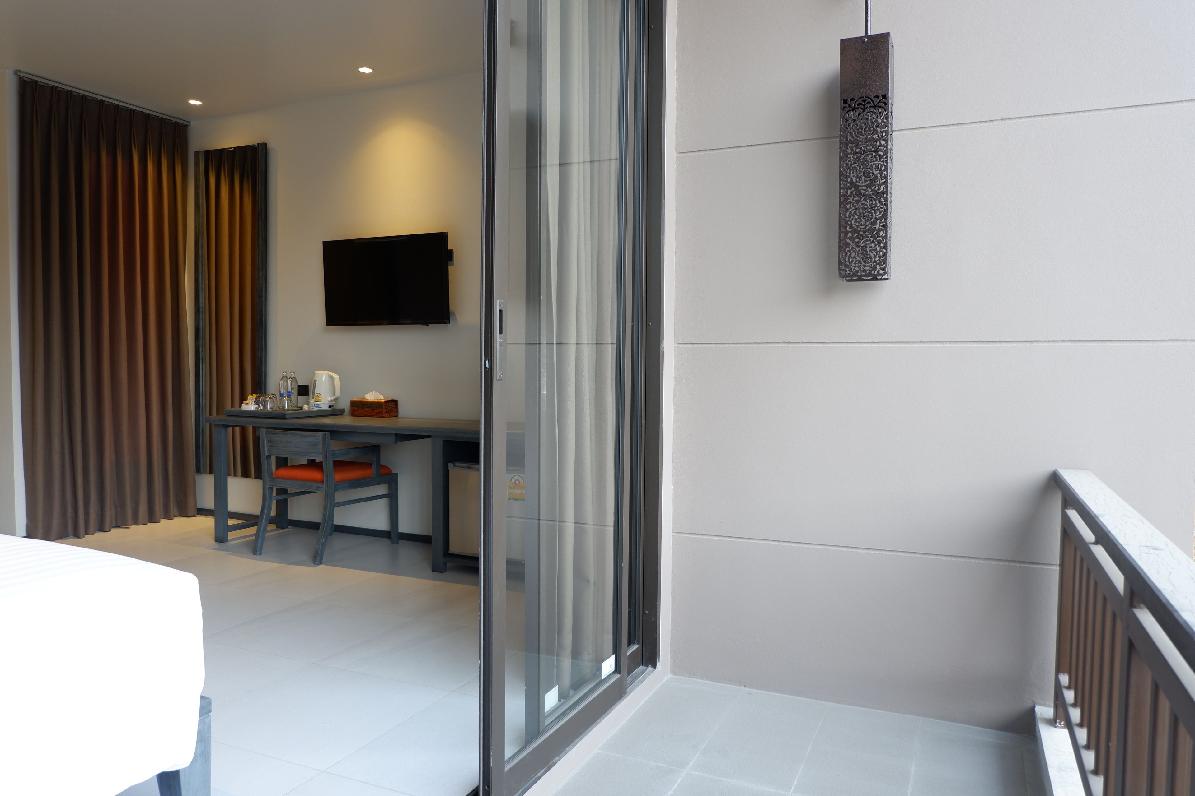 A modern hotel room with a bed, a desk with a chair, a TV mounted on the wall, and a balcony with a railing.