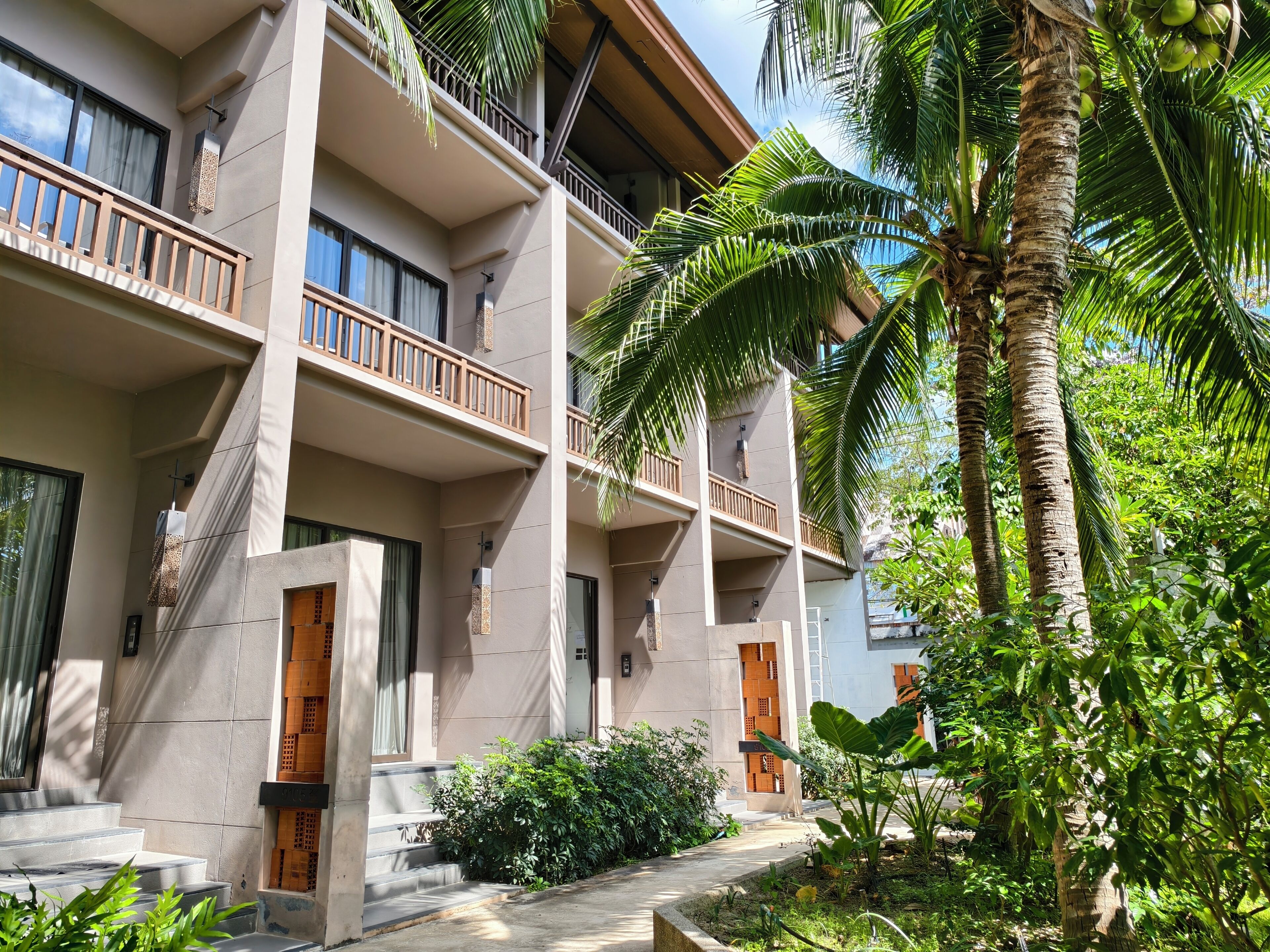 Un hôtel à plusieurs étages, doté de balcons, d’un palmier et d’un jardin bien entretenu.