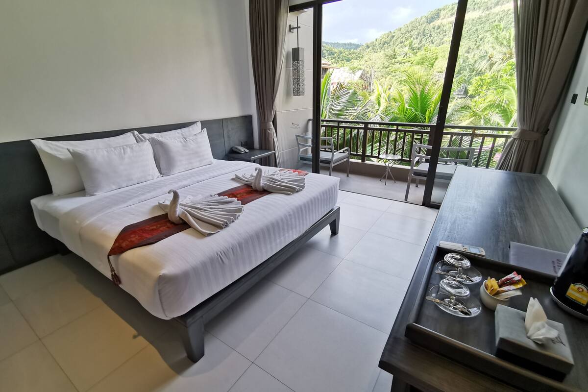 Une chambre d’hôtel avec un lit, un bureau et un balcon offrant une vue sur la verdure et les collines.