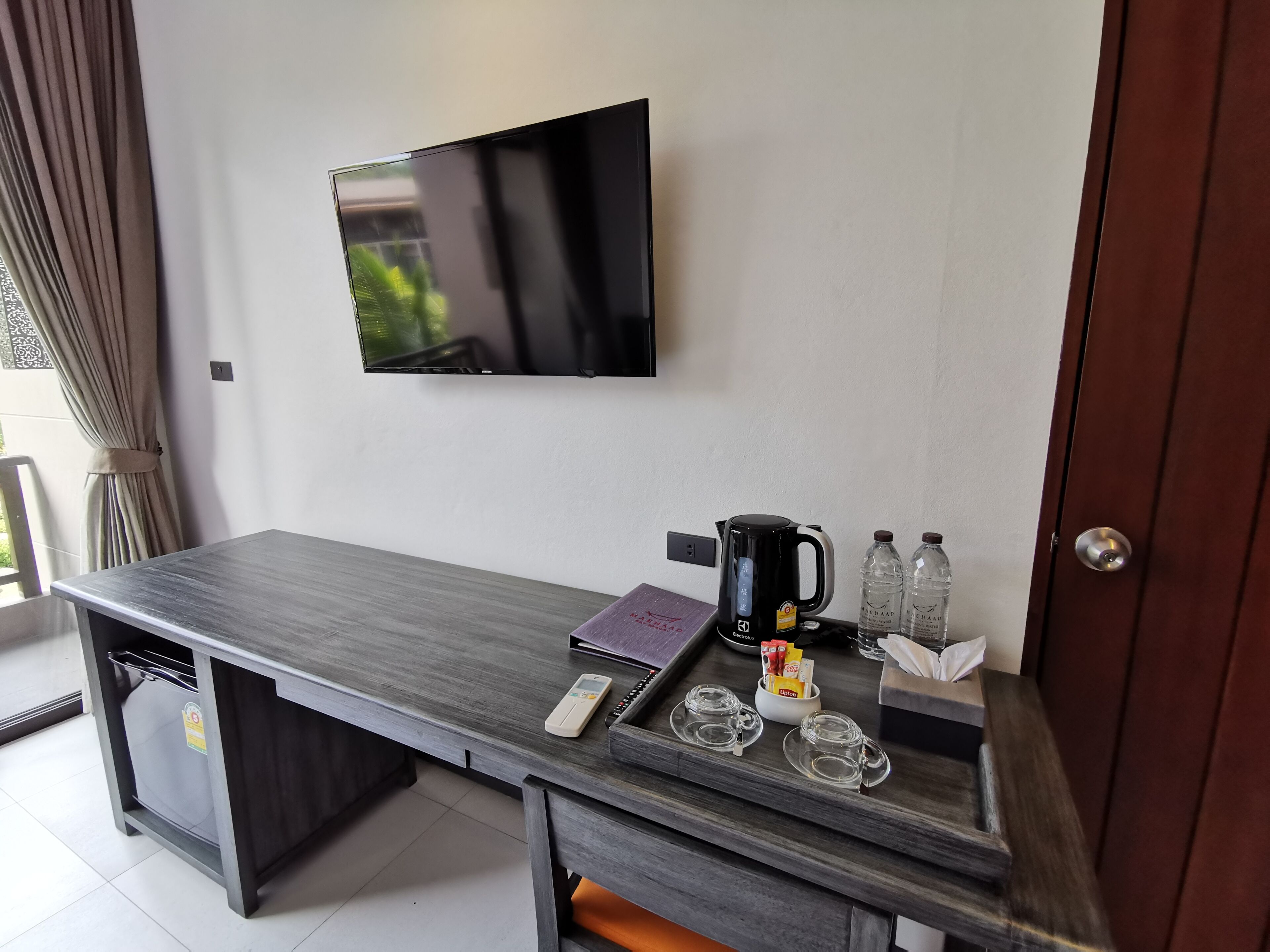 Una camera moderna con TV a schermo piatto montata a parete, una macchina per il caffè, due bottiglie d'acqua, un telecomando e un vassoio con bicchieri e scatola di fazzoletti.