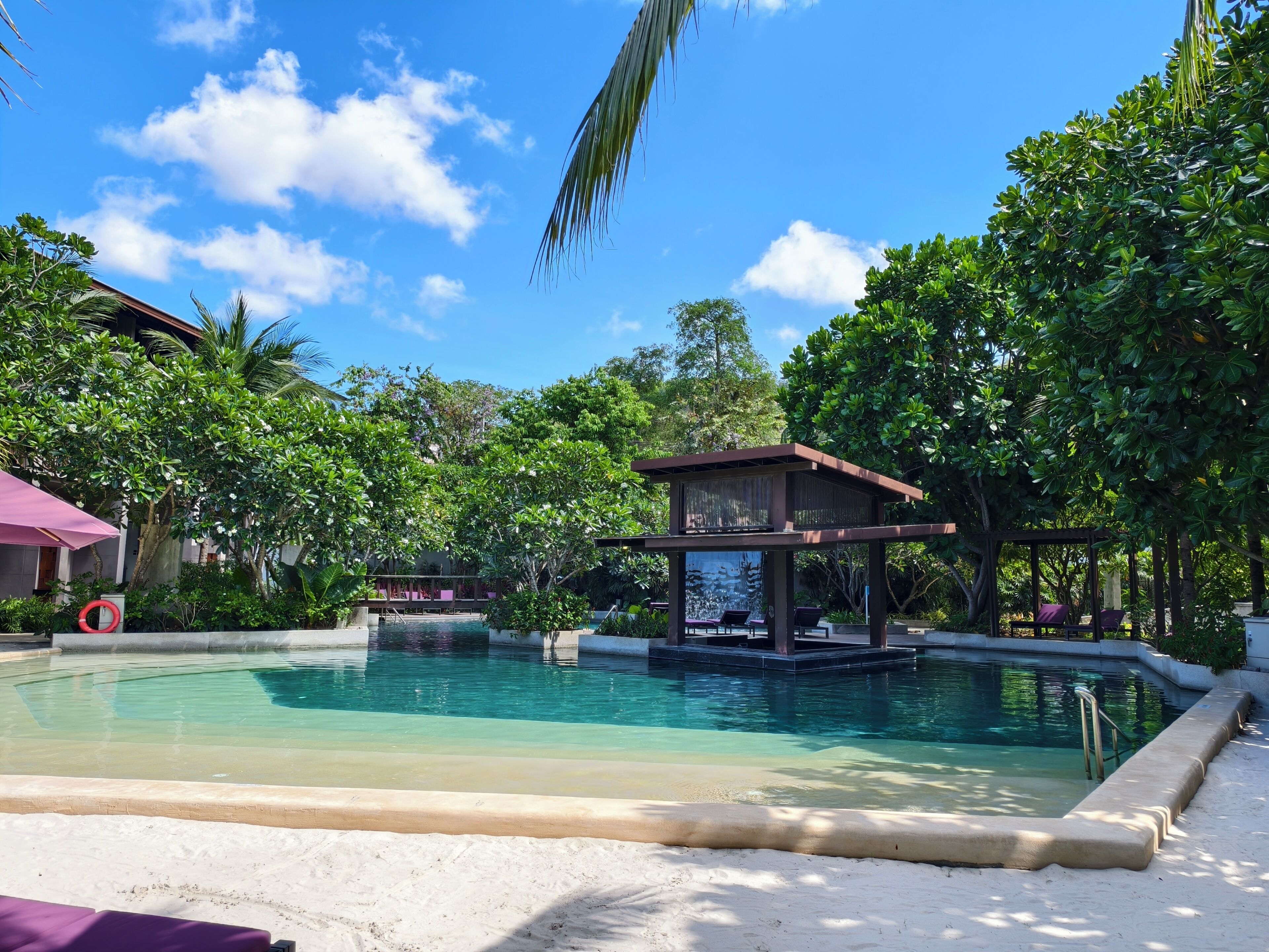 A pool area with a covered seating area, surrounded by lush greenery and a clear sky.