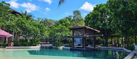 Un área de piscina con zona de estar cubierta, rodeada de vegetación exuberante y un cielo despejado.