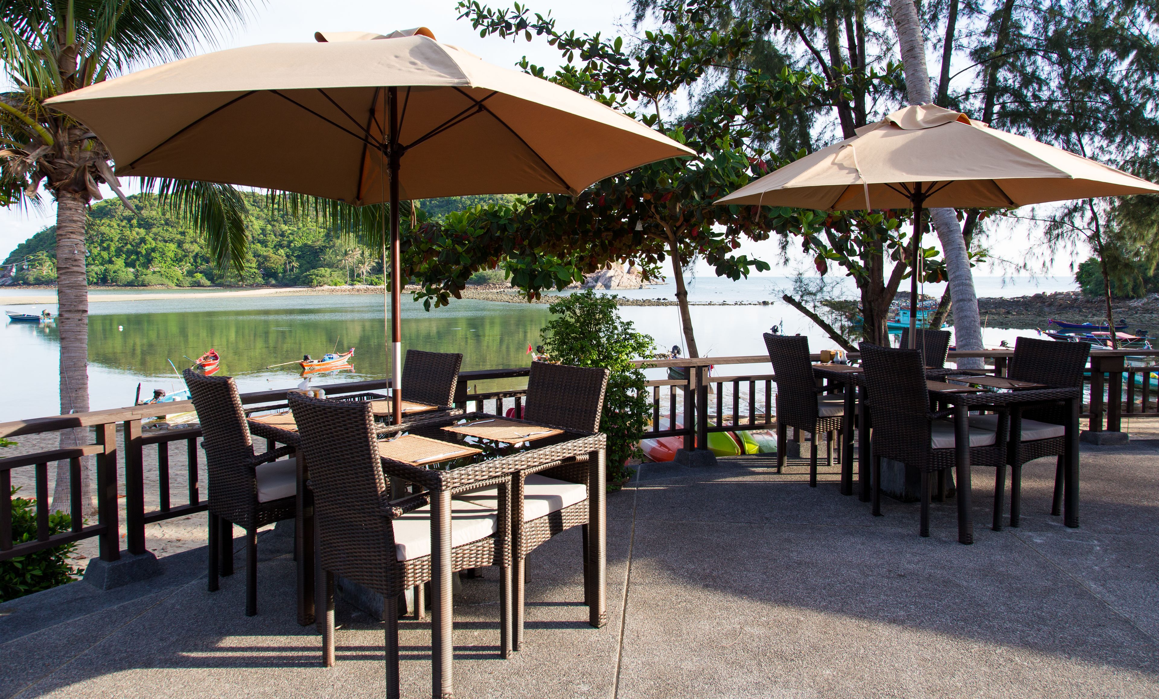 An outdoor dining area with wicker furniture and beige umbrellas, overlooking a calm lake with boats.